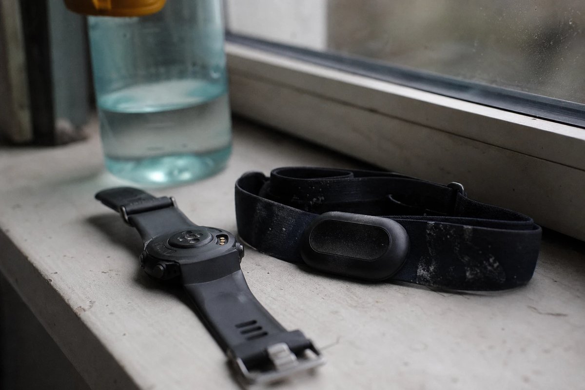 GPS-Sportuhr und ein leicht feuchter Brustgurt mit getrockneten Salzrändern auf einer Fensterbank nach einer langen Einheit, im Hintergrund eine halb leere Trinkflasche