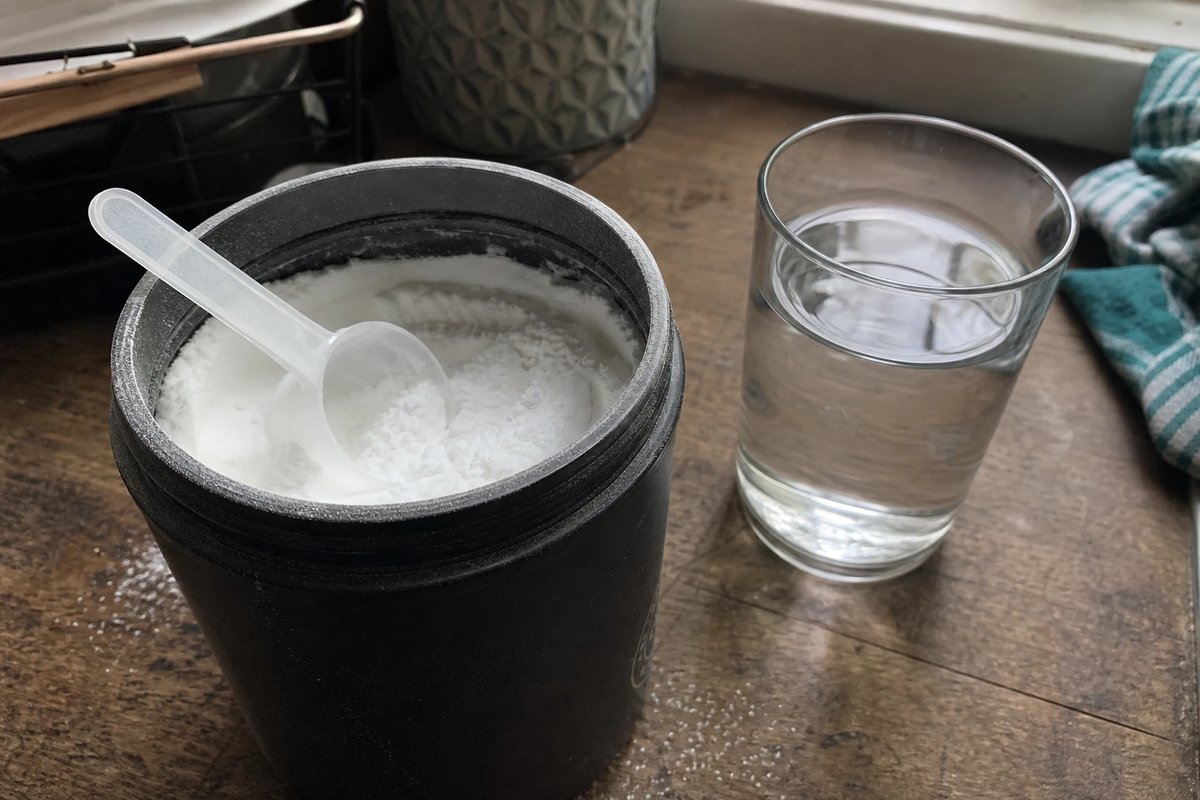 Offene Dose mit weißem Beta-Alanin-Pulver und einem kleinen Messlöffel auf einer abgenutzten Holzarbeitsplatte, daneben ein Glas Wasser