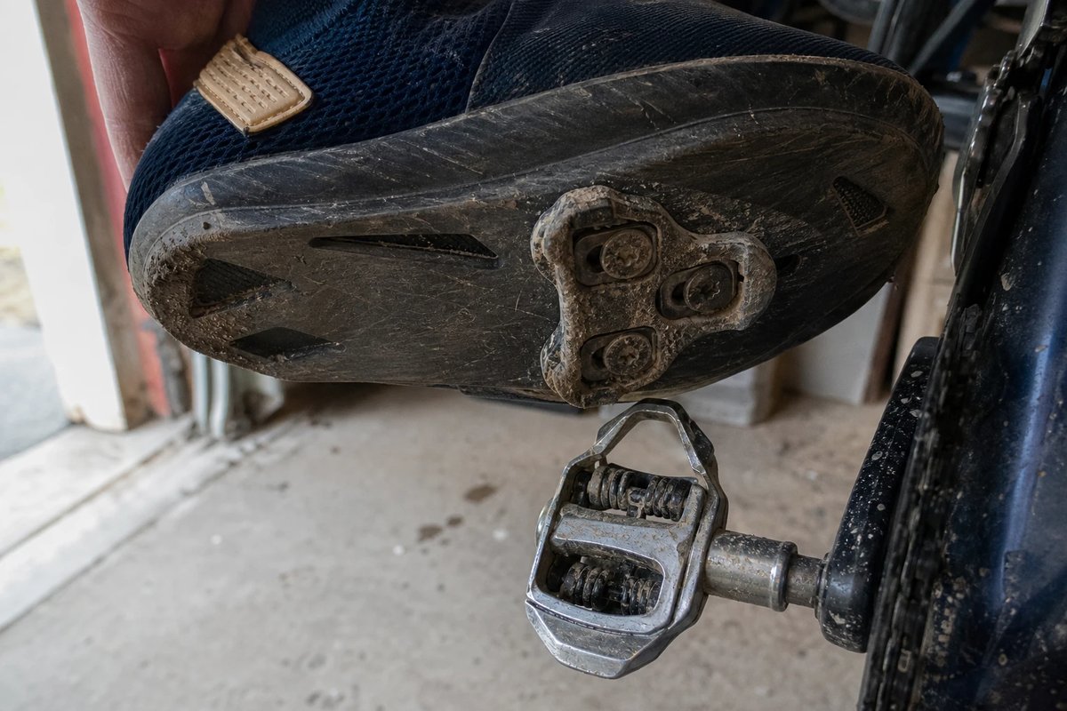 Unterseite eines abgenutzten Rennradschuhs mit einer Drei-Loch-Cleat kurz vor dem Einklicken über einem Klickpedal, leichter Staub an der Sohle