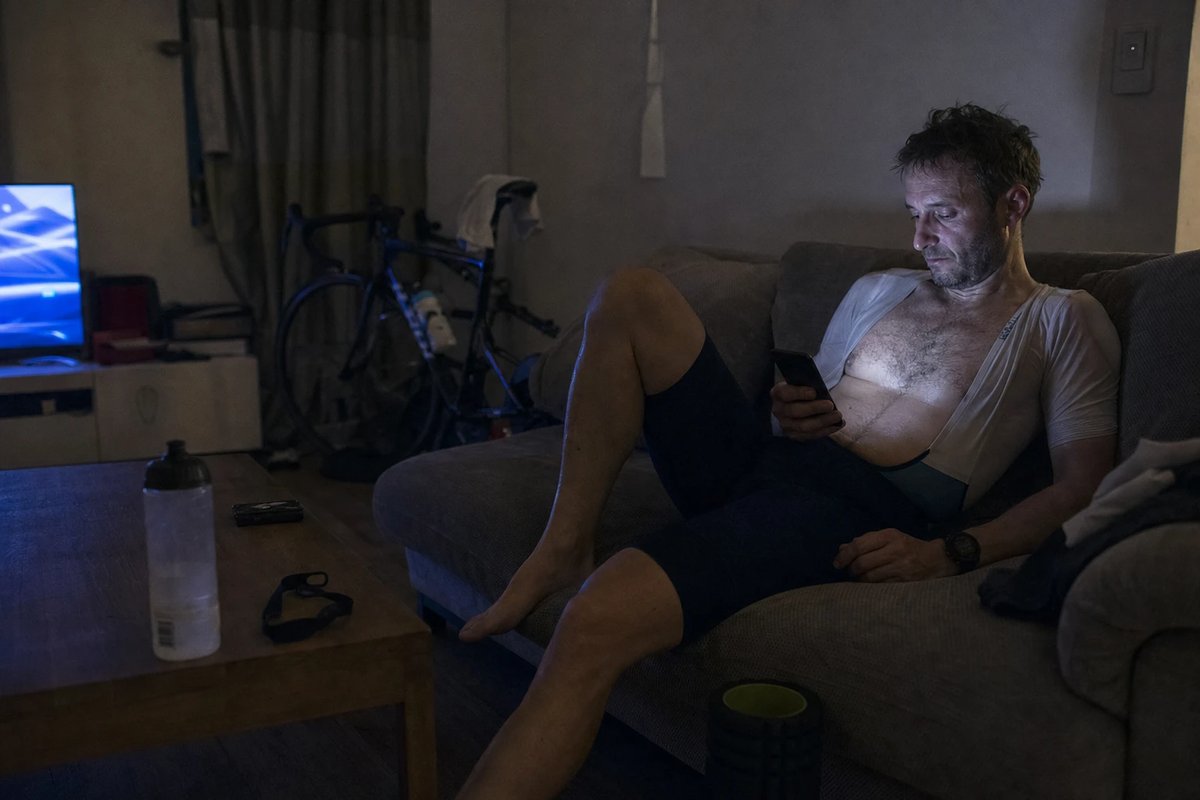 Ein müder Mann sitzt nach einer späten Smarttrainer-Einheit noch im Radtrikot auf dem Sofa und scrollt im dunklen Wohnzimmer am Handy, während im Hintergrund der Fernseher blau flackert