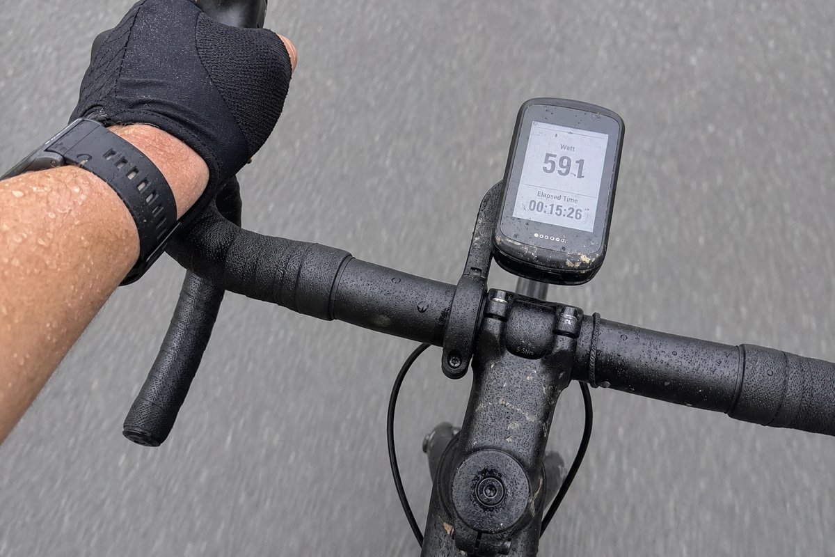 Lenkermontierter Radcomputer während einer harten Testfahrt: das Display zeigt eine hohe Wattzahl und einen laufenden Timer, ein verschwitzter Unterarm umfasst den Lenker, Schweißtropfen auf dem Oberrohr