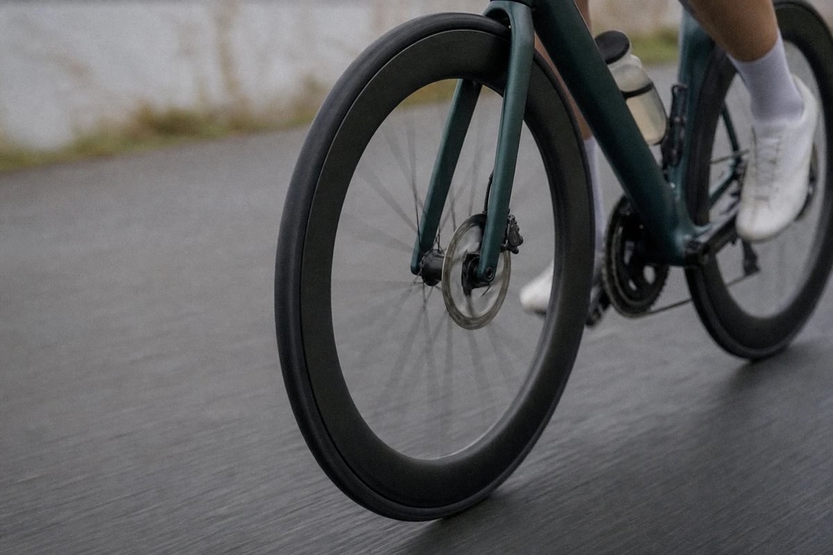 Aerodynamisches Vorderrad und schmaler Reifen eines Rennrads rollen über glatten Asphalt, leichte Bewegungsunschärfe in den Speichen, Schuh und Trinkflasche des Fahrers am oberen Bildrand