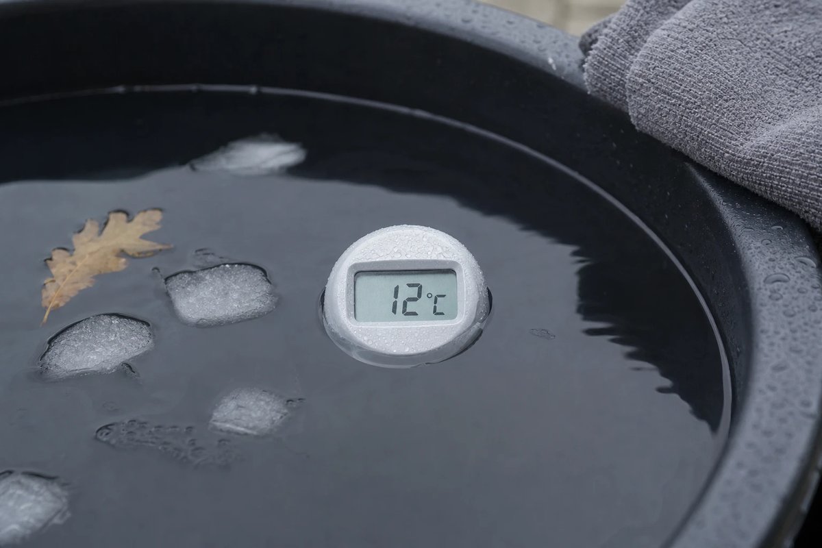 Schwimmendes digitales Thermometer in einer dunklen Eisbad-Tonne, das Display zeigt 12 Grad Celsius, daneben schmelzende Eisstücke und ein Blatt, am Rand ein graues Handtuch