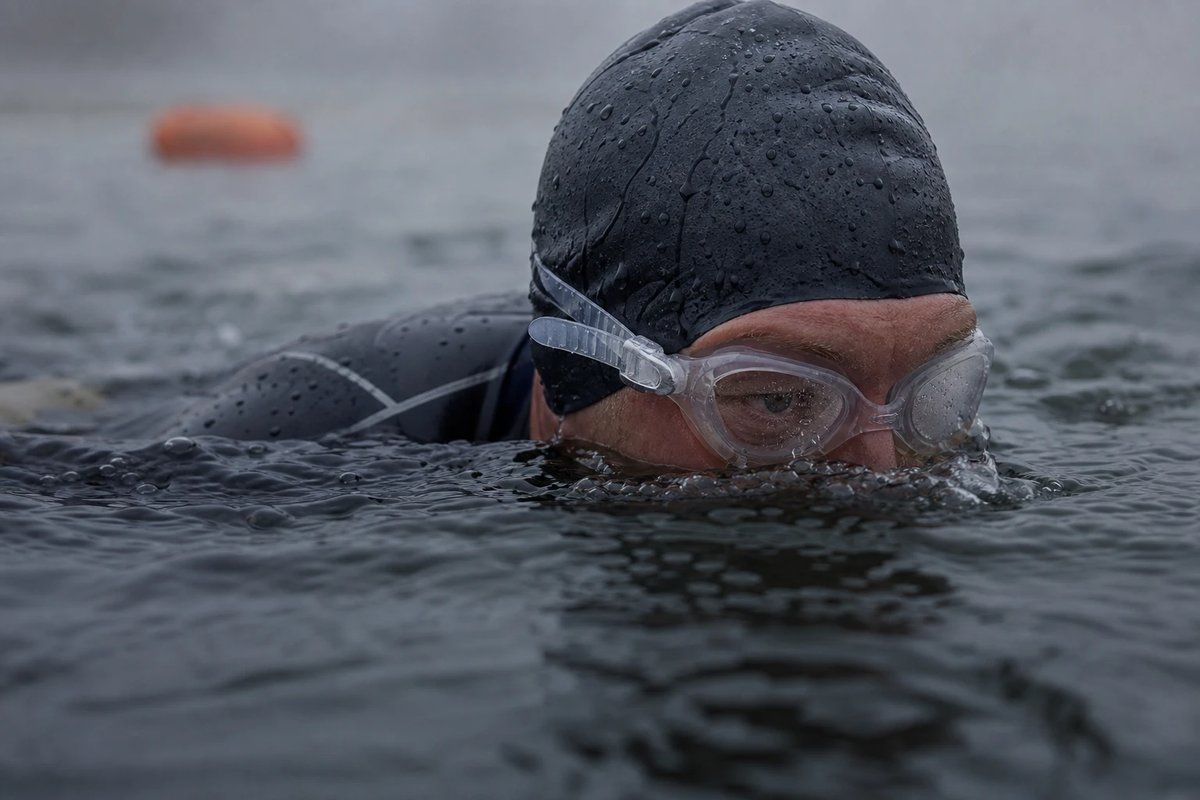 Nahaufnahme eines Freiwasserschwimmers im Neoprenanzug von der Seite an der Wasserlinie: nur die Augen und die Schwimmbrille kommen wie bei einem Krokodil über die Oberfläche, das Kinn bleibt im Wasser, im Hintergrund unscharf eine orangefarbene Boje