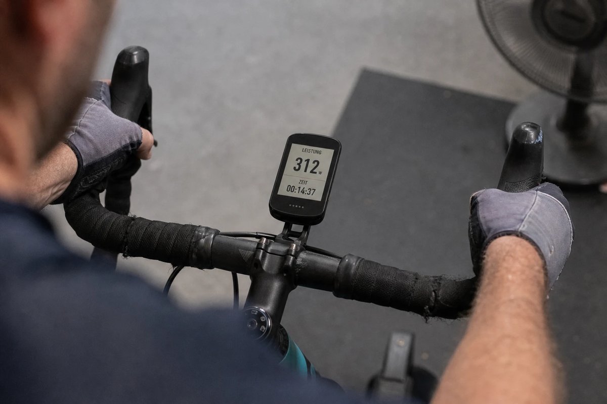 Lenkermontierter Radcomputer auf dem Smarttrainer: das Display zeigt aus Fahrersicht eine dreistellige Wattzahl und einen laufenden Timer, behandschuhte Hände am Lenker