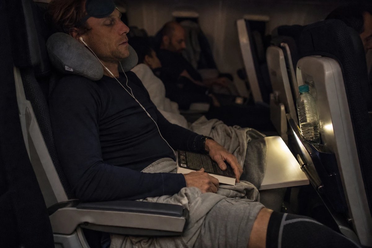Ausdauerathlet schläft im engen Economy-Sitz auf einem Nachtflug, Schlafmaske auf der Stirn, Nackenkissen und dunkle Kompressionsstrümpfe, Wasserflasche in der Sitztasche