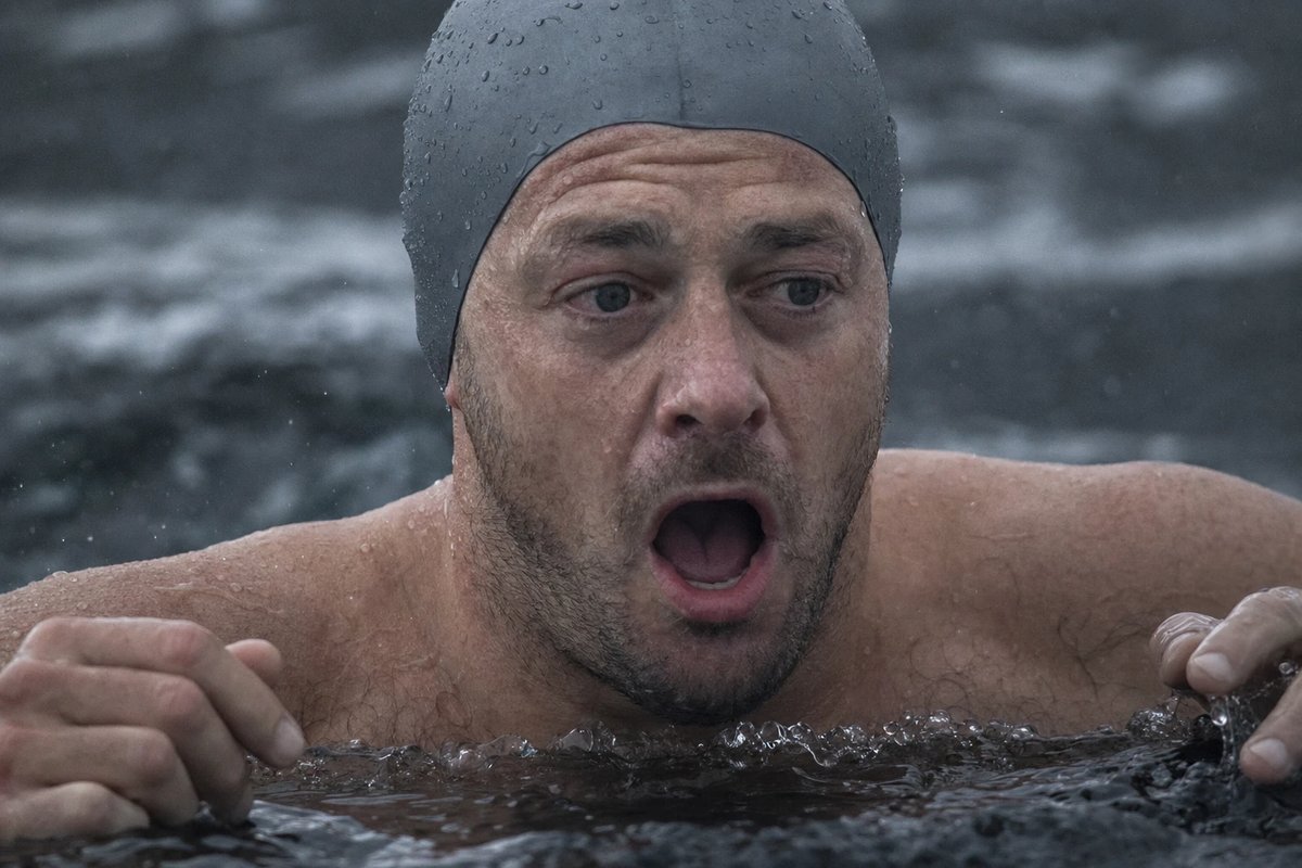 Ein Freiwasserschwimmer mit Badekappe taucht brusttief in dunkles, kaltes Wasser ein und schnappt unwillkürlich nach Luft, das Gesicht von Wassertropfen bedeckt