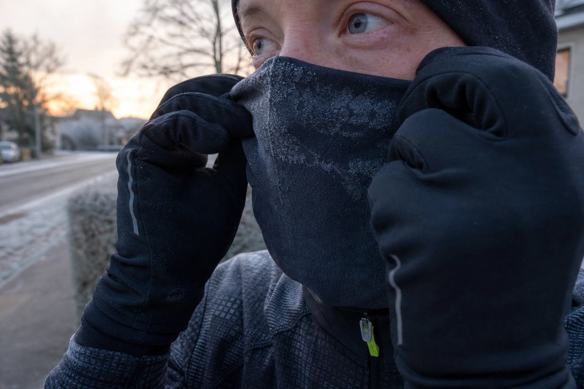 Läuferin zieht sich vor einem Lauf im frostigen Morgengrauen mit behandschuhten Händen ein Multifunktionstuch über Mund und Nase, im Hintergrund eine vereiste Straße