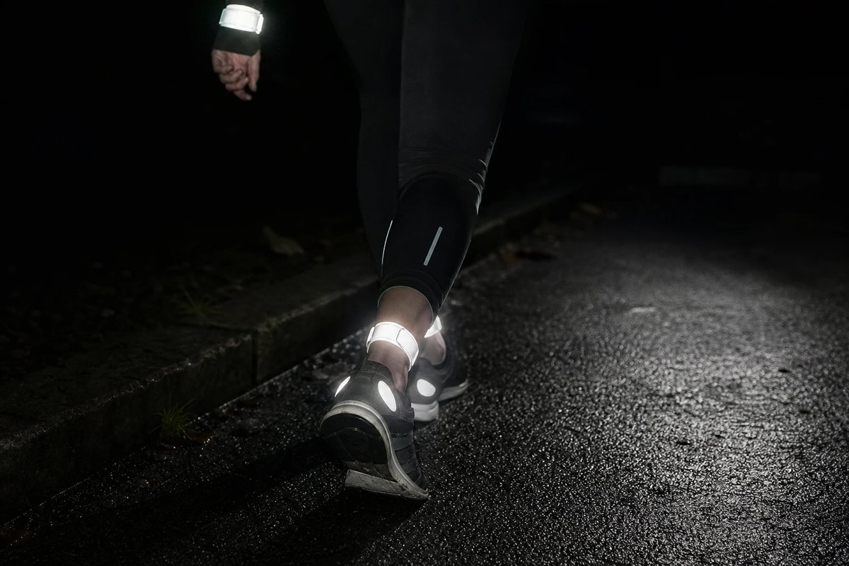 Nahaufnahme der Unterschenkel und Knöchel einer Läuferin bei Nacht, von einem Autoscheinwerfer seitlich angestrahlt, sodass die Reflektorstreifen an beiden Knöcheln und an den Schuhfersen hell aufleuchten, während alles andere im Dunkeln liegt