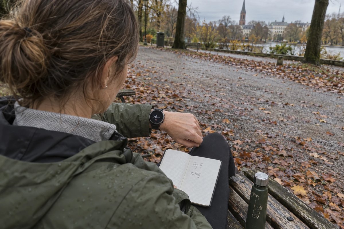 Frau auf einer Parkbank nach einem kurzen Spaziergang, von hinten über die Schulter fotografiert, ihre Sportuhr zeigt 96 Schläge pro Minute, auf dem Knie ein Notizbuch mit den Worten Tag 4 und ruhig