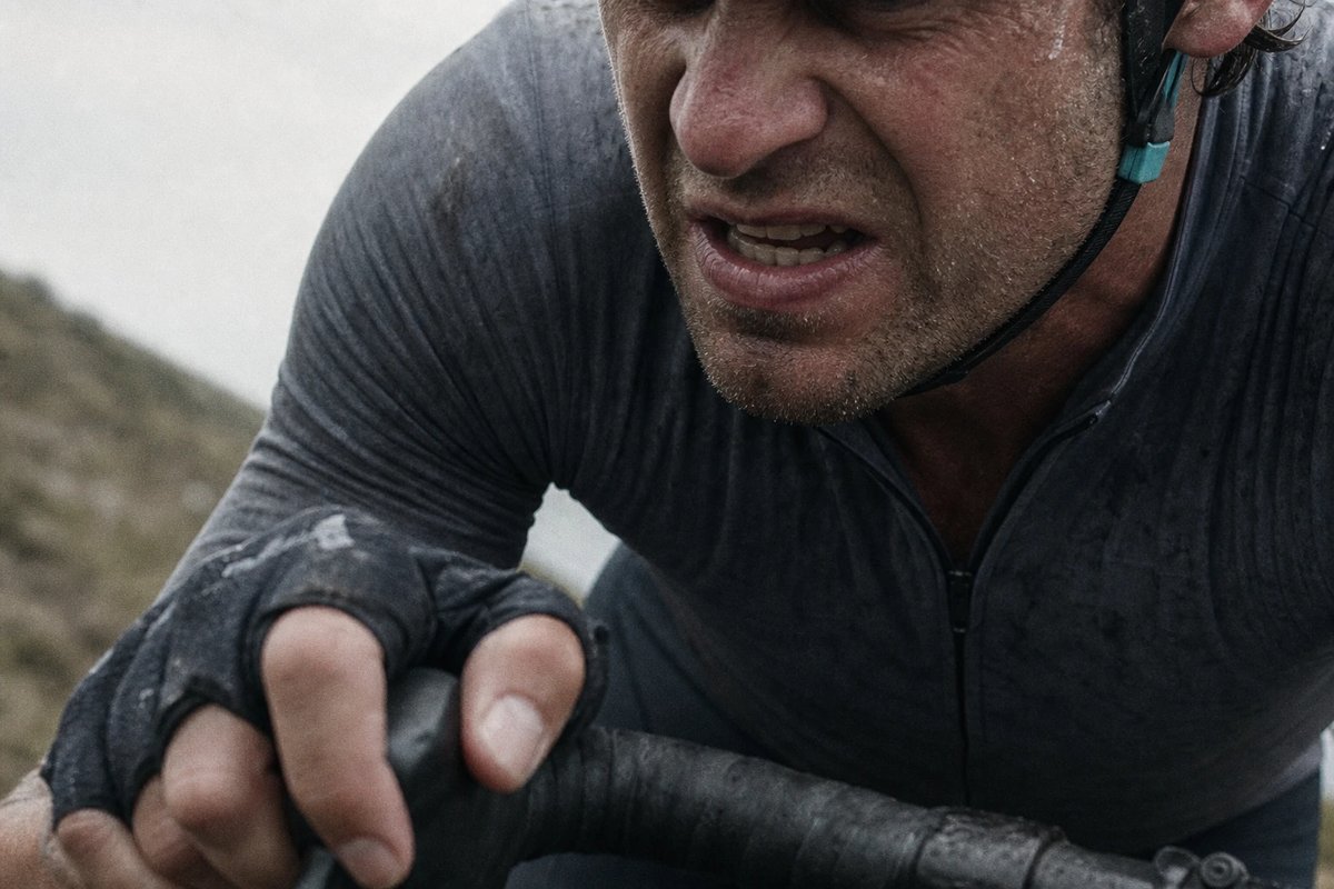 Nahaufnahme des angestrengten Gesichts eines Rennradfahrers an einem Anstieg, Kiefer angespannt und Schweiß an der Schläfe, eine behandschuhte Hand am Lenkerband