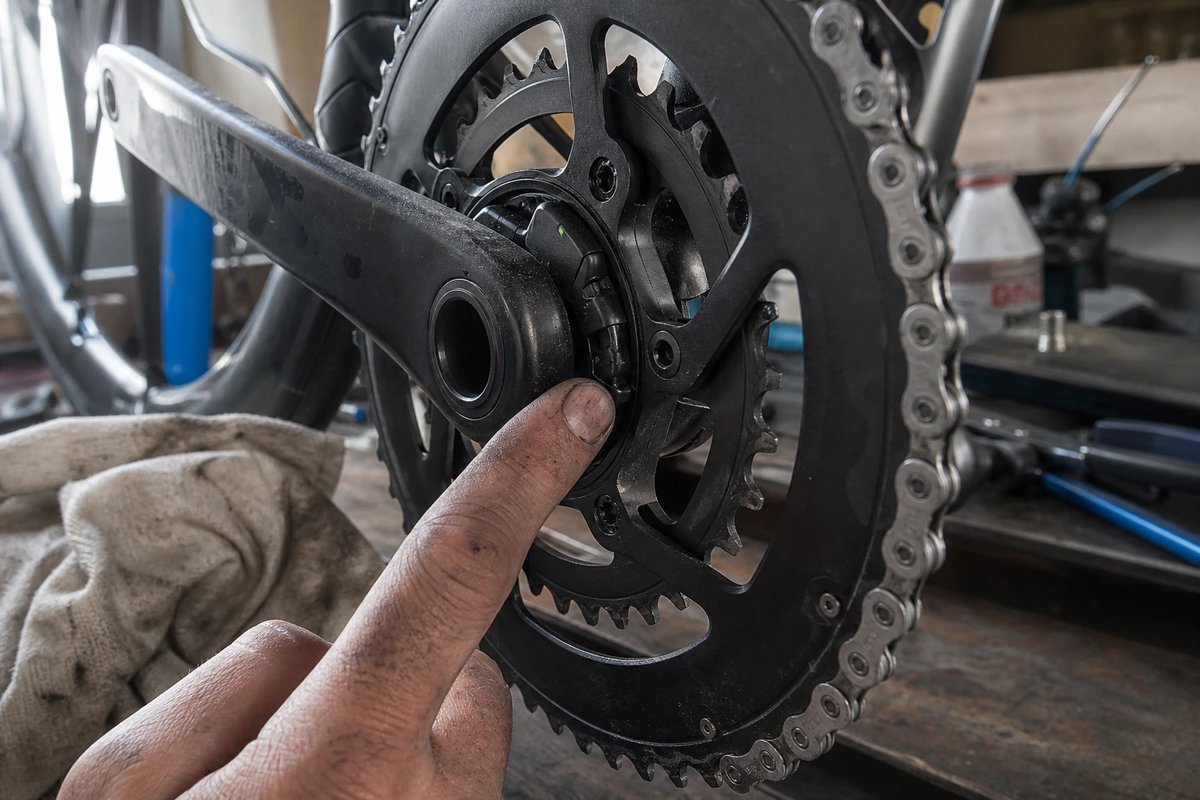 Nahaufnahme eines Spider-Powermeters im Kettenblattträger einer Rennrad-Kurbel, ein Finger mit Kettenfett zeigt auf die kleine Sensoreinheit zwischen Kurbelarm und Kettenblatt, im Hintergrund eine unscharfe Werkbank