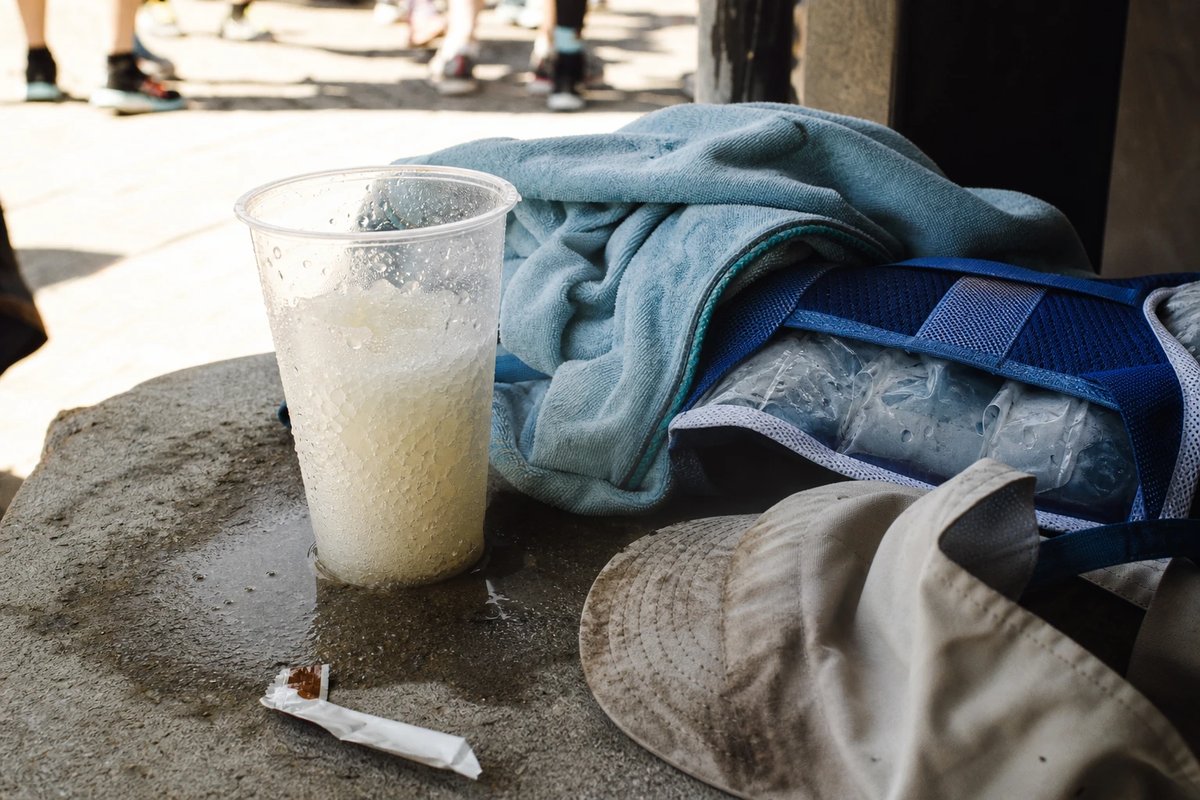 Schattige Bank an einem heißen Wettkampfstart mit einem halb geschmolzenen Becher Eis-Slurry, einem feuchten Kühlhandtuch und einer zusammengelegten Kühlweste mit sichtbaren Kühlpacks, Kondenswasser am Becher