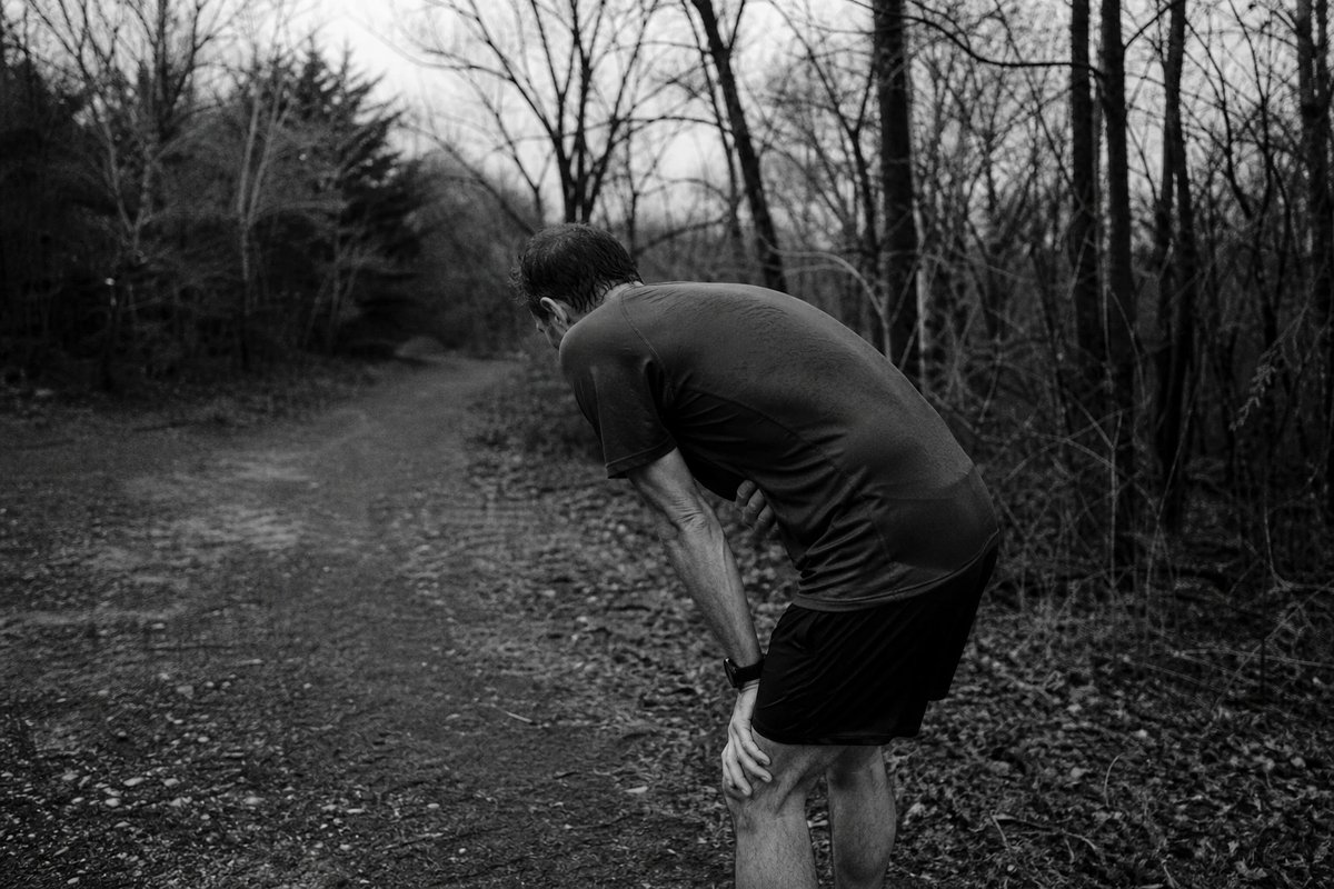 Amateurläufer steht auf einem Waldweg, vornübergebeugt mit den Händen auf den Knien, eine Hand am Bauch, sichtlich von Magen-Darm-Beschwerden ausgebremst