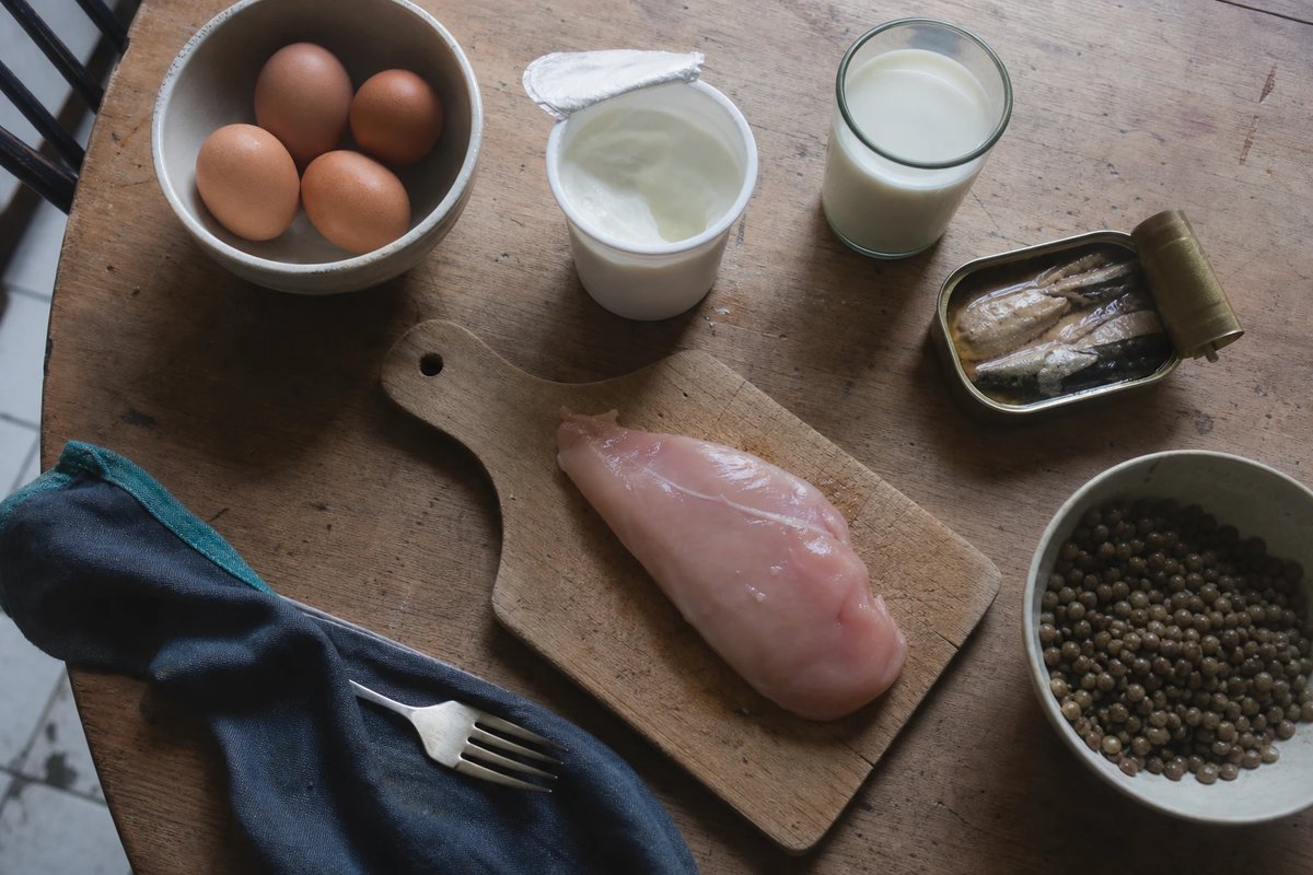 Aufsicht auf einen Holztisch mit alltäglichen Proteinquellen: eine Schale Eier, ein offener Becher Quark, ein Glas Milch, eine Dose Fisch, eine rohe Hähnchenbrust und eine Schale gekochte Linsen