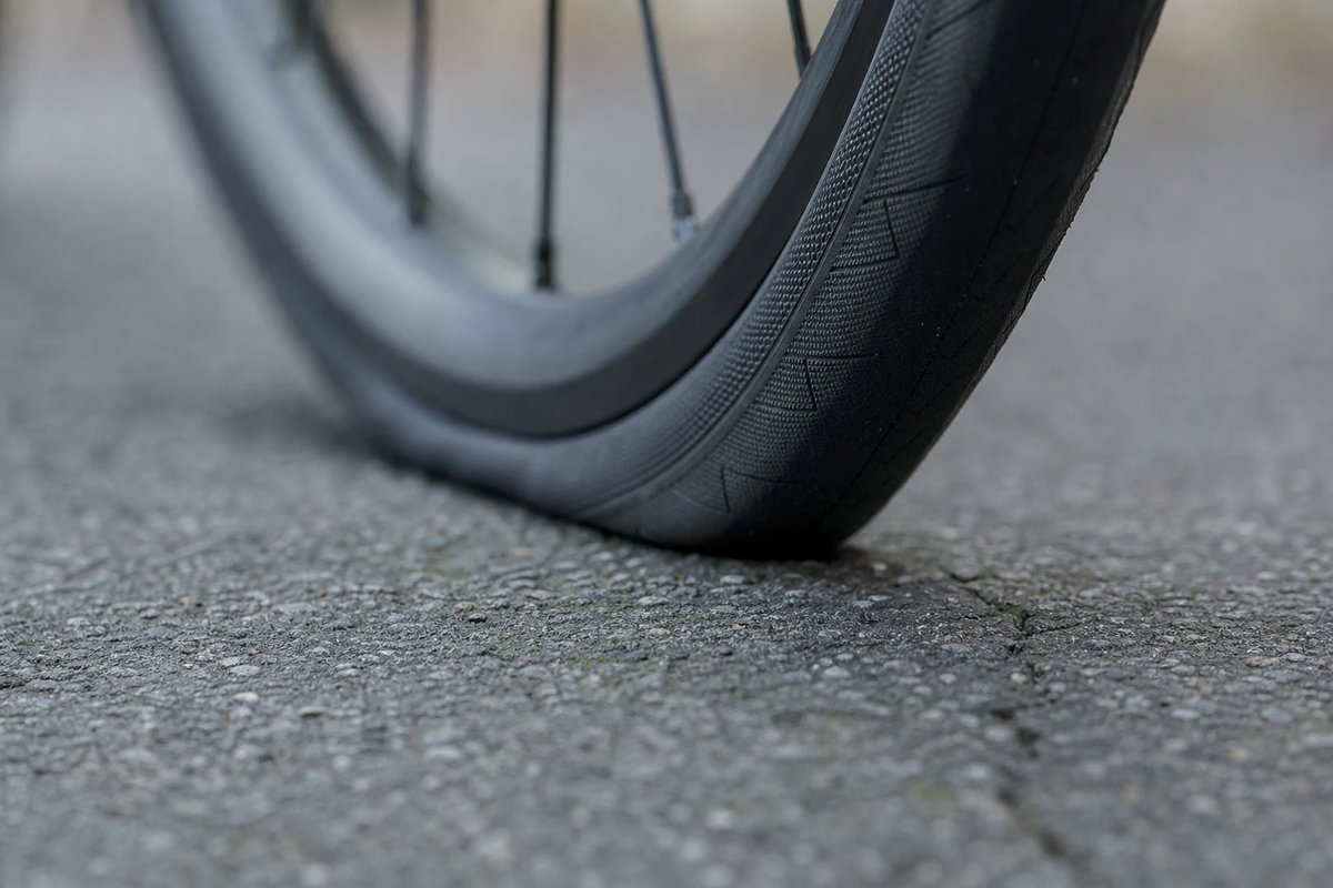 Nahaufnahme eines Rennradreifens auf rauem grauem Asphalt: die kurze Aufstandsfläche und die feine Reifenverformung dort, wo der Gummi den Boden berührt