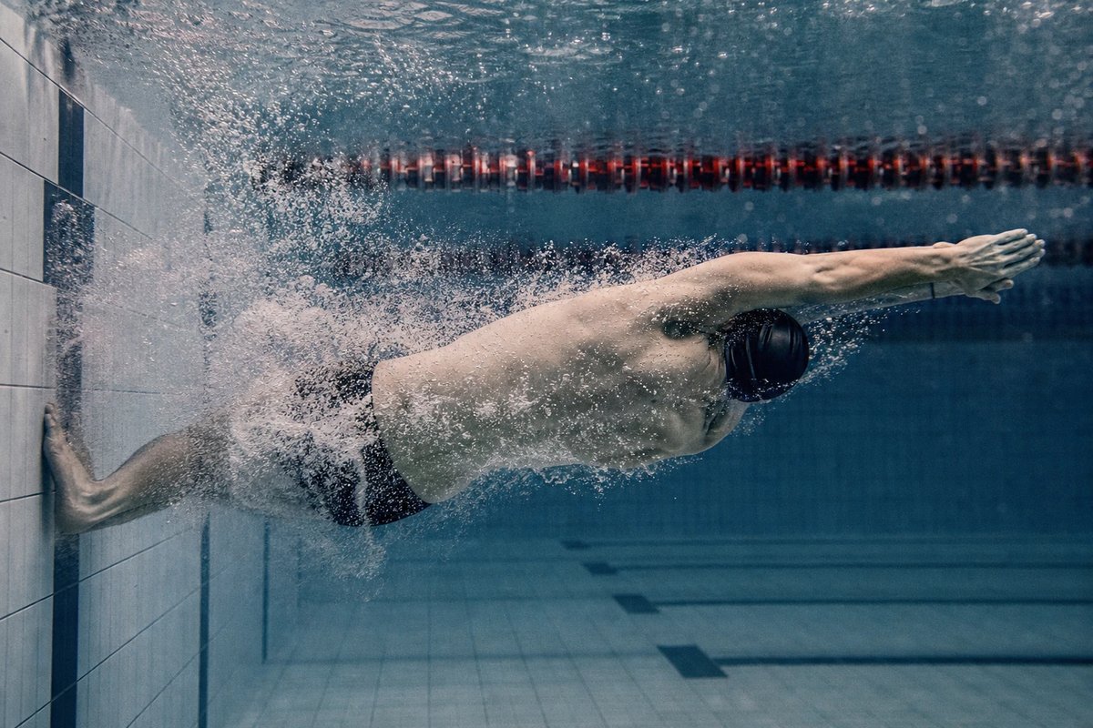 Unterwasseraufnahme eines Schwimmers im Abdruckmoment der Rollwende: beide Füße an der Wand, Beine strecken sich kraftvoll, Hände übereinander über dem Kopf, Arme an die Ohren gepresst, ein dichter Blasenschweif folgt dem Körper