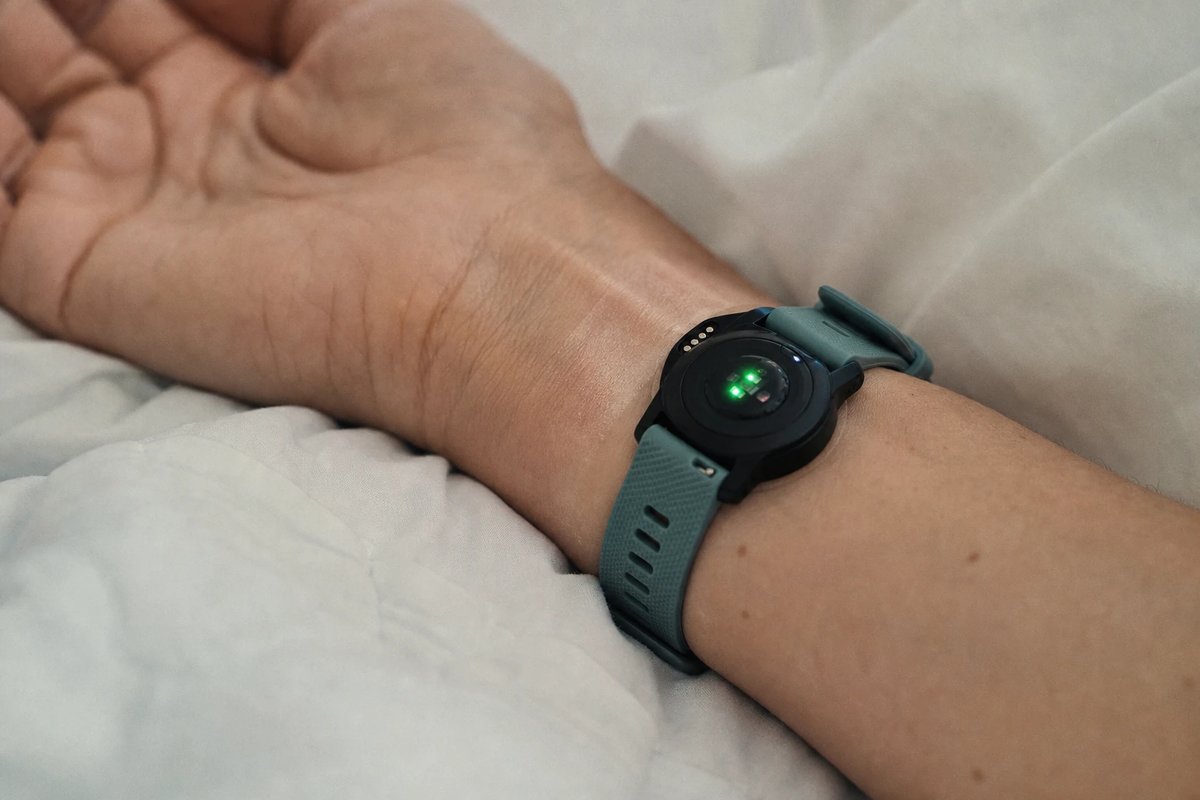 Handgelenk auf einer weißen Bettdecke am frühen Morgen, an der Unterseite einer Fitnessuhr leuchtet der grüne optische Herzfrequenzsensor gegen die Haut