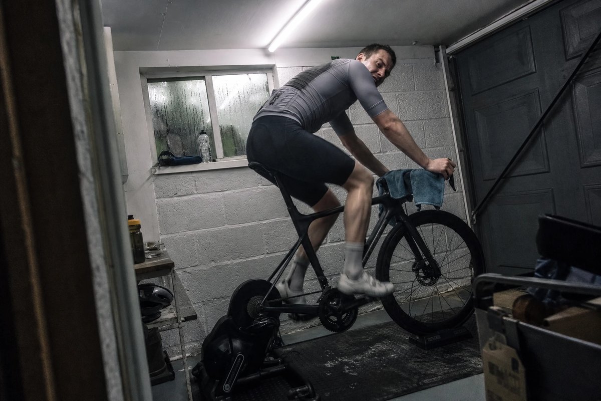 Freizeitradfahrer im maximalen Sprint auf einem Rennrad, das auf einem Smarttrainer in einer Garage eingespannt ist, mit Ventilator und schweißnassem Handtuch über dem Lenker