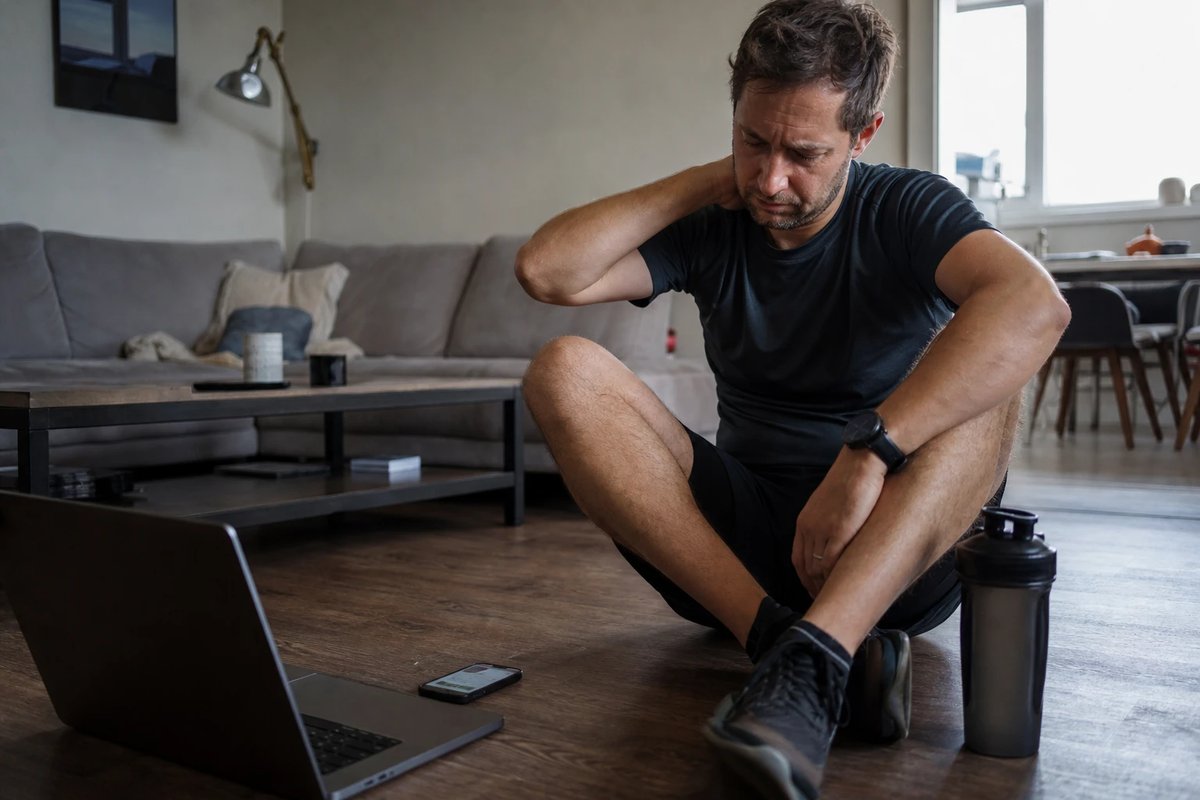 Ein Mann in Sportkleidung sitzt erschöpft und gestresst auf dem Wohnzimmerboden, reibt sich den Nacken, daneben ein Laptop und ein Handy mit Benachrichtigungen