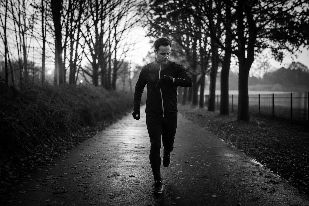 Läufer auf einem kurzen, lockeren Dauerlauf auf einem ruhigen baumgesäumten Weg im ersten Tageslicht, Blick auf die GPS-Uhr zur Tempokontrolle