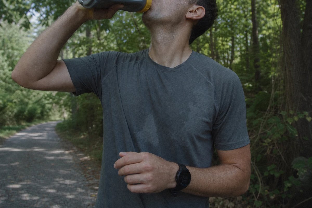 Läufer pausiert mitten in einem langen Lauf auf einem schattigen Weg und trinkt aus einer Softflask, das Shirt schweißfeucht, eine GPS-Uhr am Handgelenk