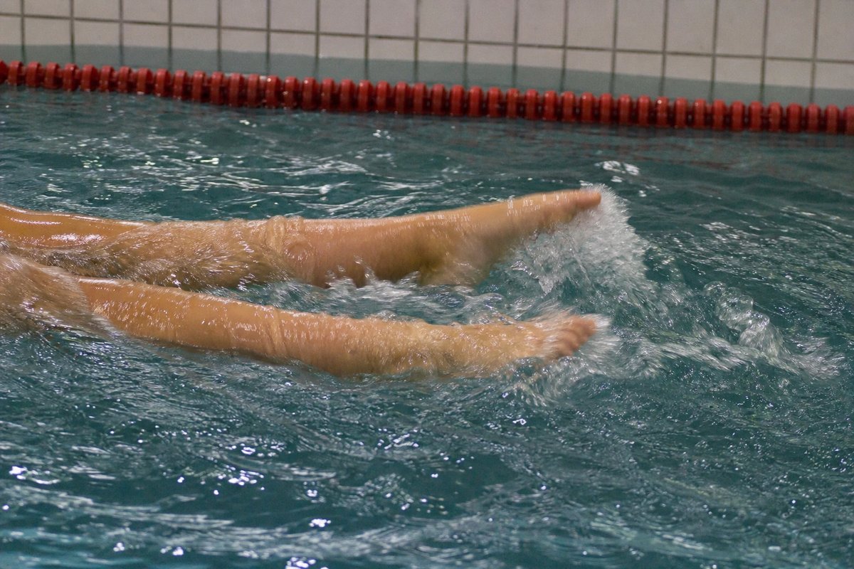 Nahaufnahme der Füße und Unterschenkel einer Schwimmerin mitten im Flutter Kick an der Wasseroberfläche: lockere Sprunggelenke, gestreckte Füße in leichter Plantarflexion, eine niedrige Schaumspur an den Zehen, oben im Bild eine rote Leinenboje