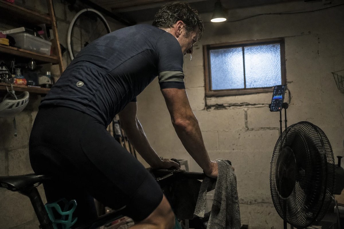 Rennradfahrer im Wiegetritt bei einem harten VO2max-Intervall auf einem Smarttrainer in einem Keller-Paincave, daneben ein Ventilator und ein Display mit dem Workout