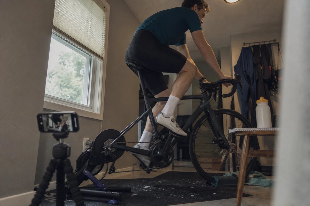 Amateur-Radfahrer tritt auf einem an einen Rollentrainer montierten Rennrad in die Pedale, während ein Handy auf einem niedrigen Stativ das Bein von der Seite filmt