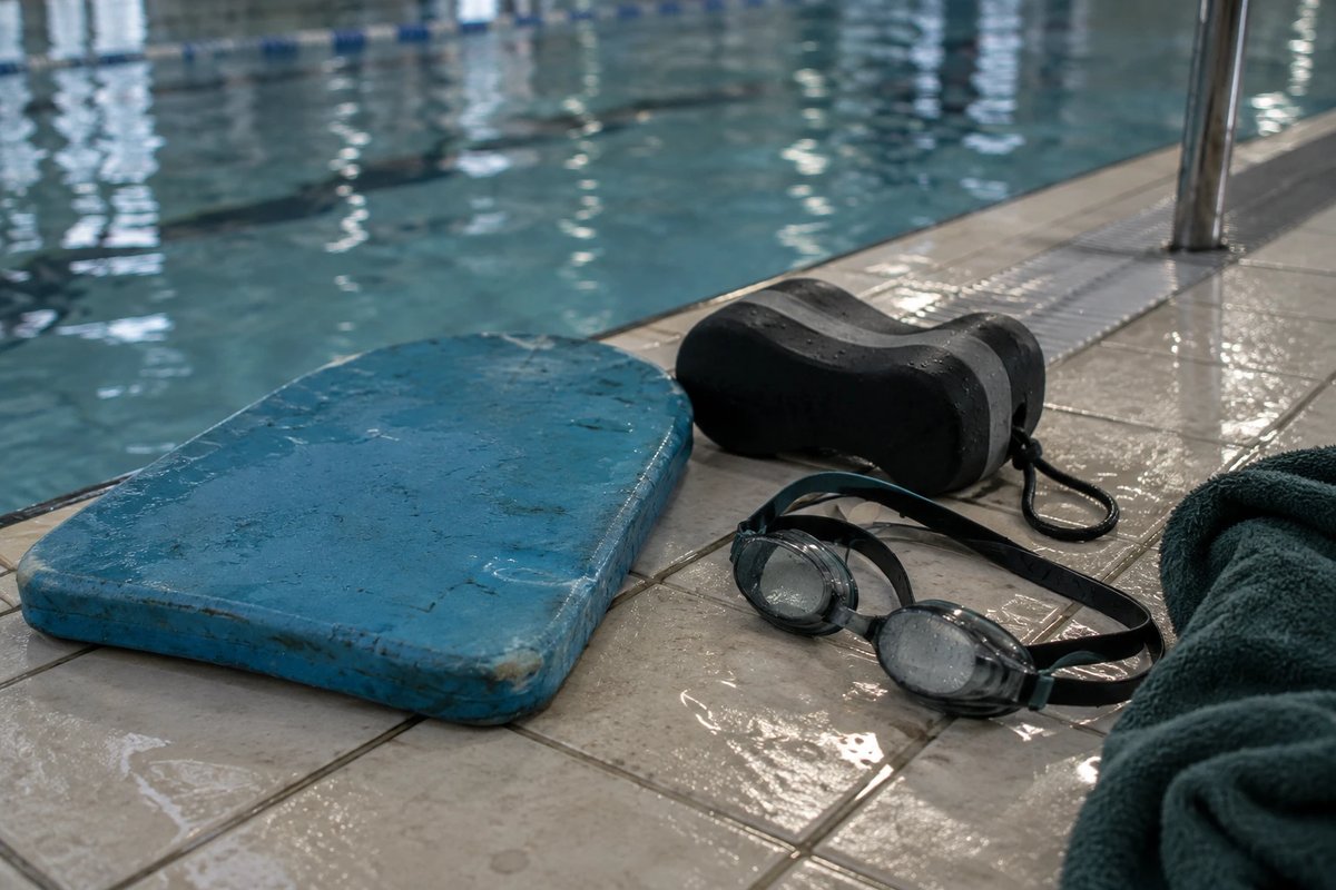 Abgenutztes blaues Schwimmbrett, schwarzer Pull Buoy, beschlagene Schwimmbrille und ein dunkelgrünes Handtuch auf nassen Fliesen am Rand eines Hallenbads nach einer Technikeinheit