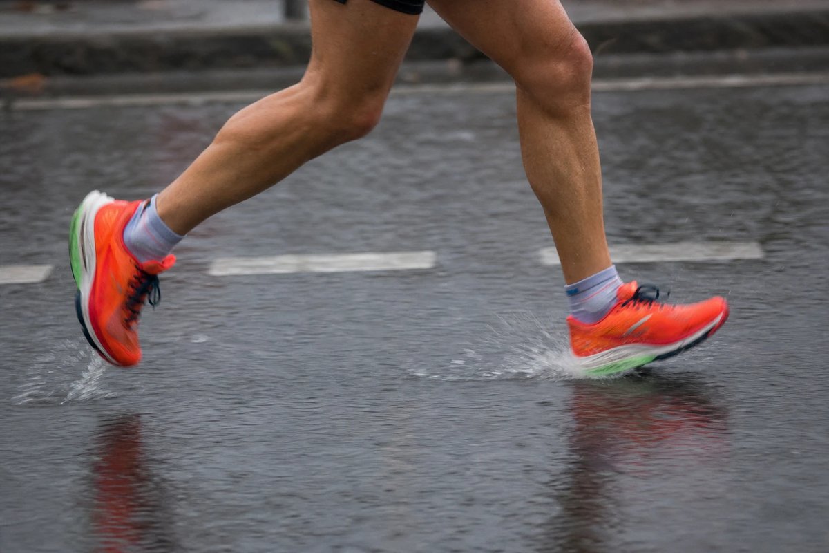 Unterschenkel und leuchtende Wettkampfschuhe eines Läufers im Laufschritt auf nasser Straße, Wasser spritzt unter den Sohlen auf