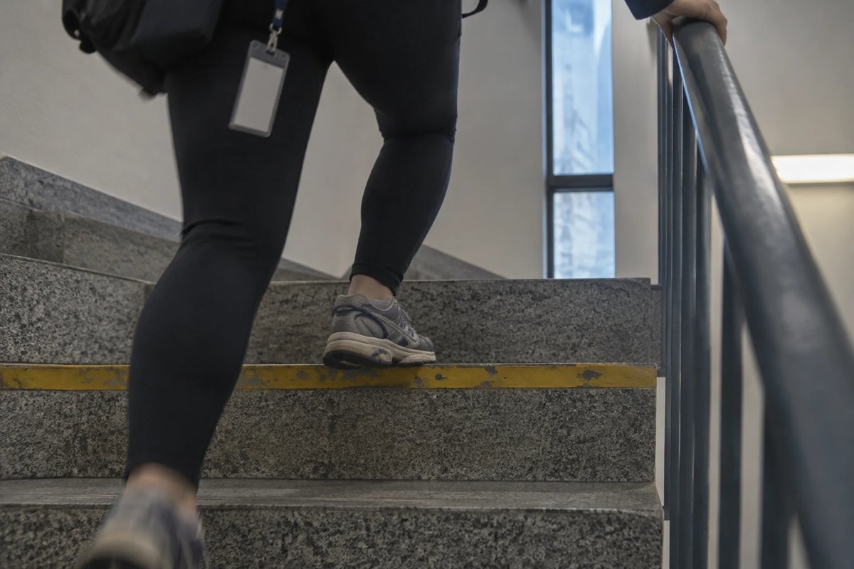 Beine in dunkler Laufhose und abgetragenen Laufschuhen auf den Stufen eines Bürotreppenhauses mit gelber Trittkante, eine Hand am Geländer, am Rand ein Mitarbeiterausweis an einem Band