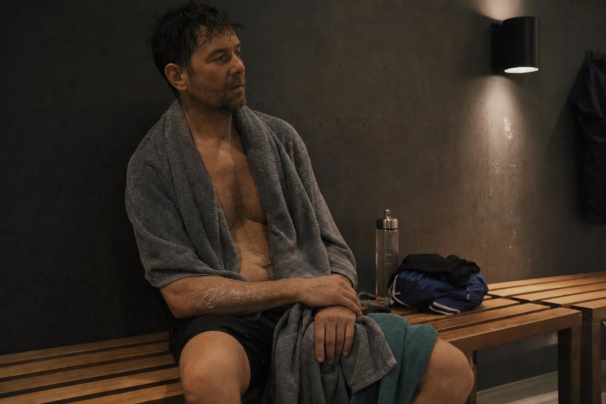 Sportler sitzt nach einer Floating-Sitzung ruhig und entspannt in ein Handtuch gewickelt auf einer Holzbank im gedämpften Licht eines Float-Studios, daneben eine Trinkflasche und zusammengelegte Laufkleidung
