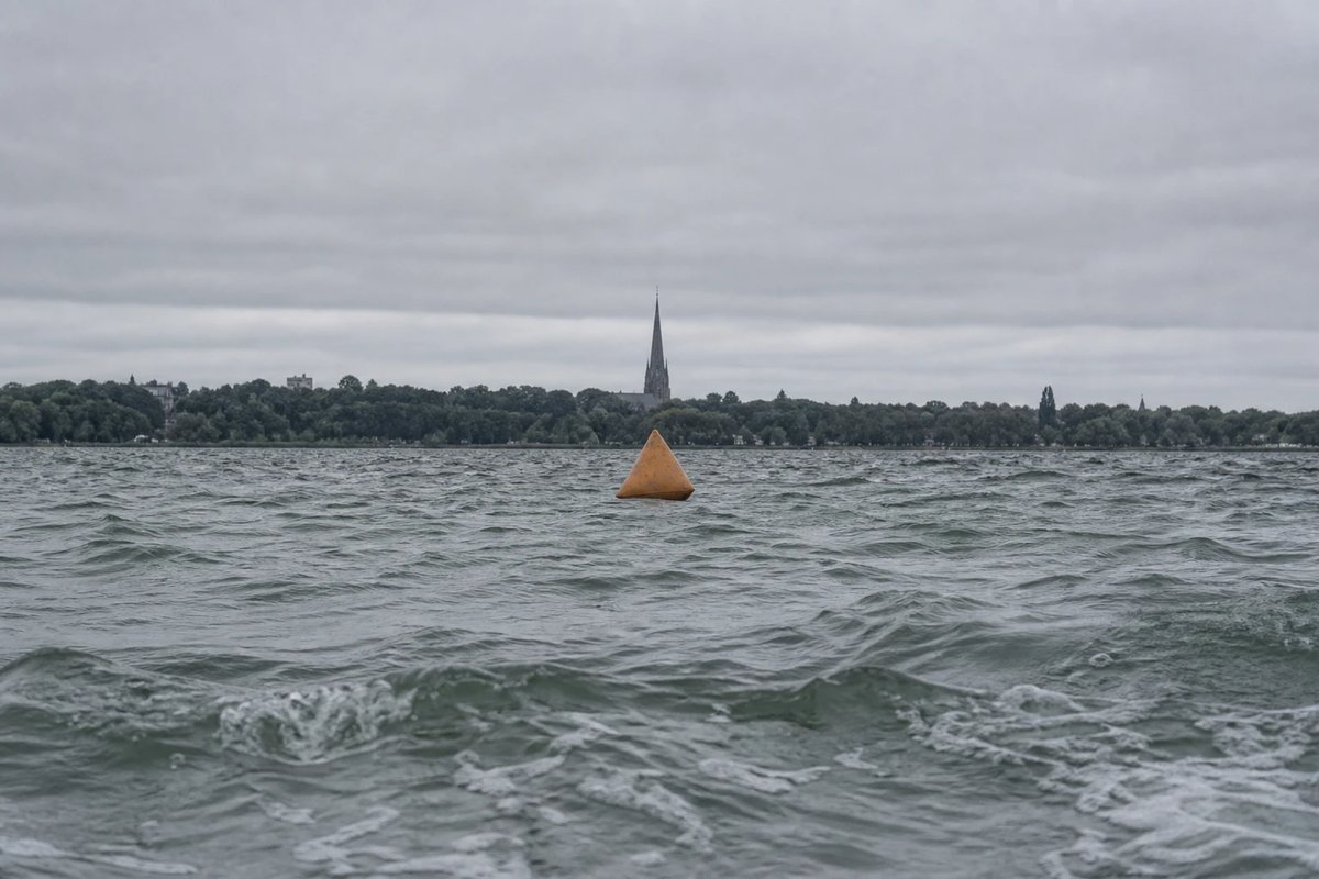 Blick flach über einen welligen grauen See aus Schwimmerperspektive: eine orangefarbene dreieckige Boje, dahinter am Horizont in einer Linie ein hoher Kirchturm als feste Landmarke zum Anpeilen