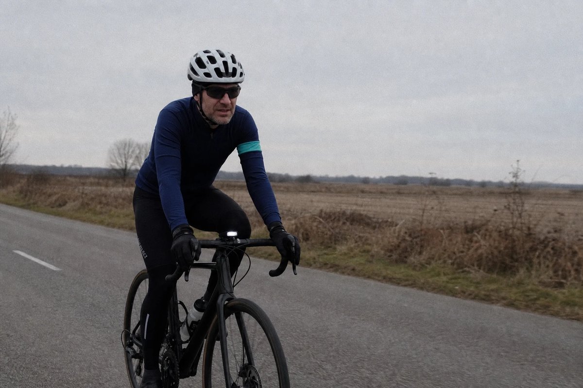 Radfahrer in lockerer, aufrechter Haltung bei ruhigem Grundlagentempo auf einer flachen grauen Landstraße an einem bedeckten Morgen