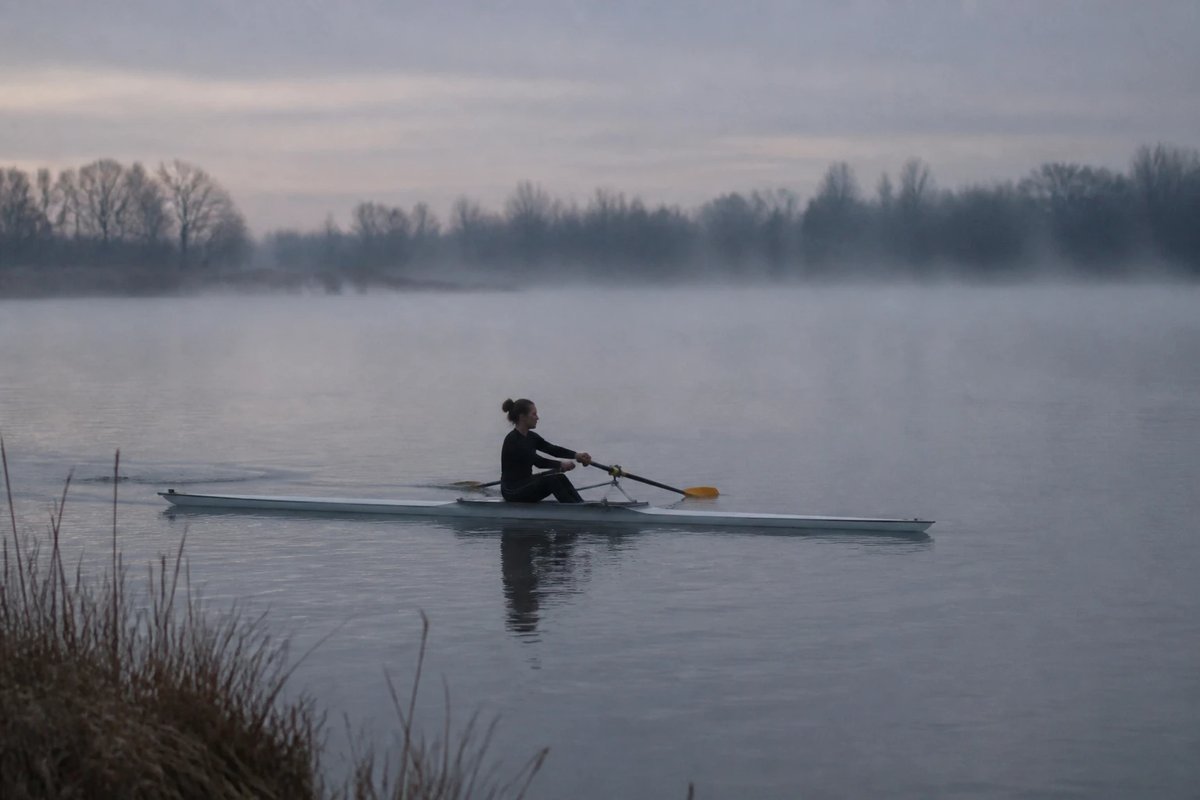 Eine einzelne Ruderin in einem Einer auf einem stillen, nebligen See im Morgengrauen, das schmale Boot und die langen Skulls liegen flach auf dem grauen Wasser