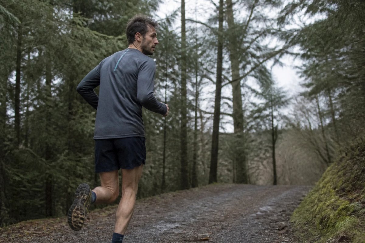 Läufer in schlichter grauer und dunkelblauer Sportkleidung auf einem nassen Waldweg an einem bedeckten Morgen, mittlere Anstrengung, von schräg hinten aufgenommen