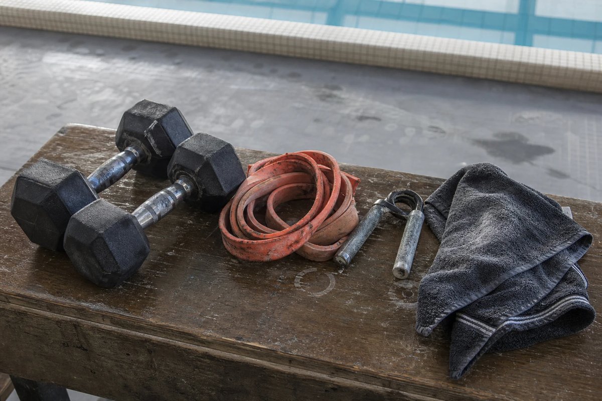 Hanteln, ein zusammengerolltes Widerstandsband, ein Handtrainer und ein feuchtes Handtuch nebeneinander auf einer Holzbank in einer Trainingsecke am Beckenrand