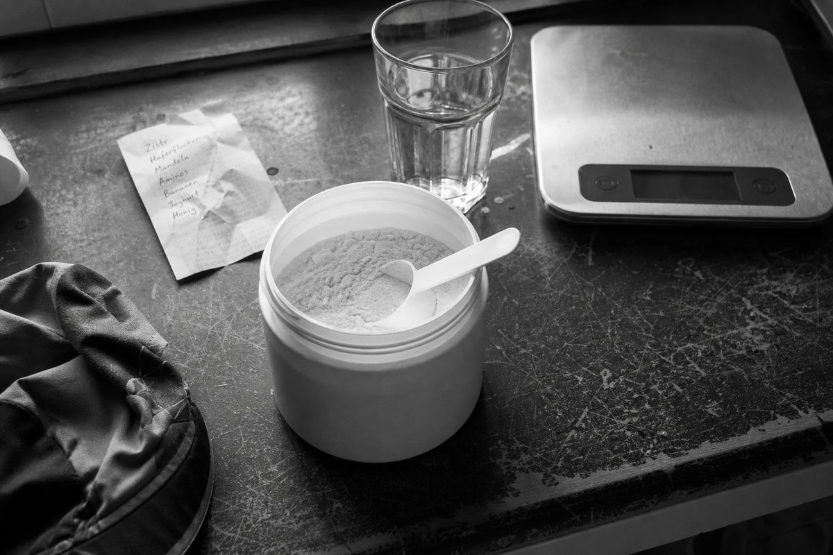 Offene Dose mit weißem Aminosäure-Pulver, Messlöffel, ein Glas Wasser und eine Küchenwaage auf einer abgenutzten Arbeitsplatte, Schwarz-Weiß
