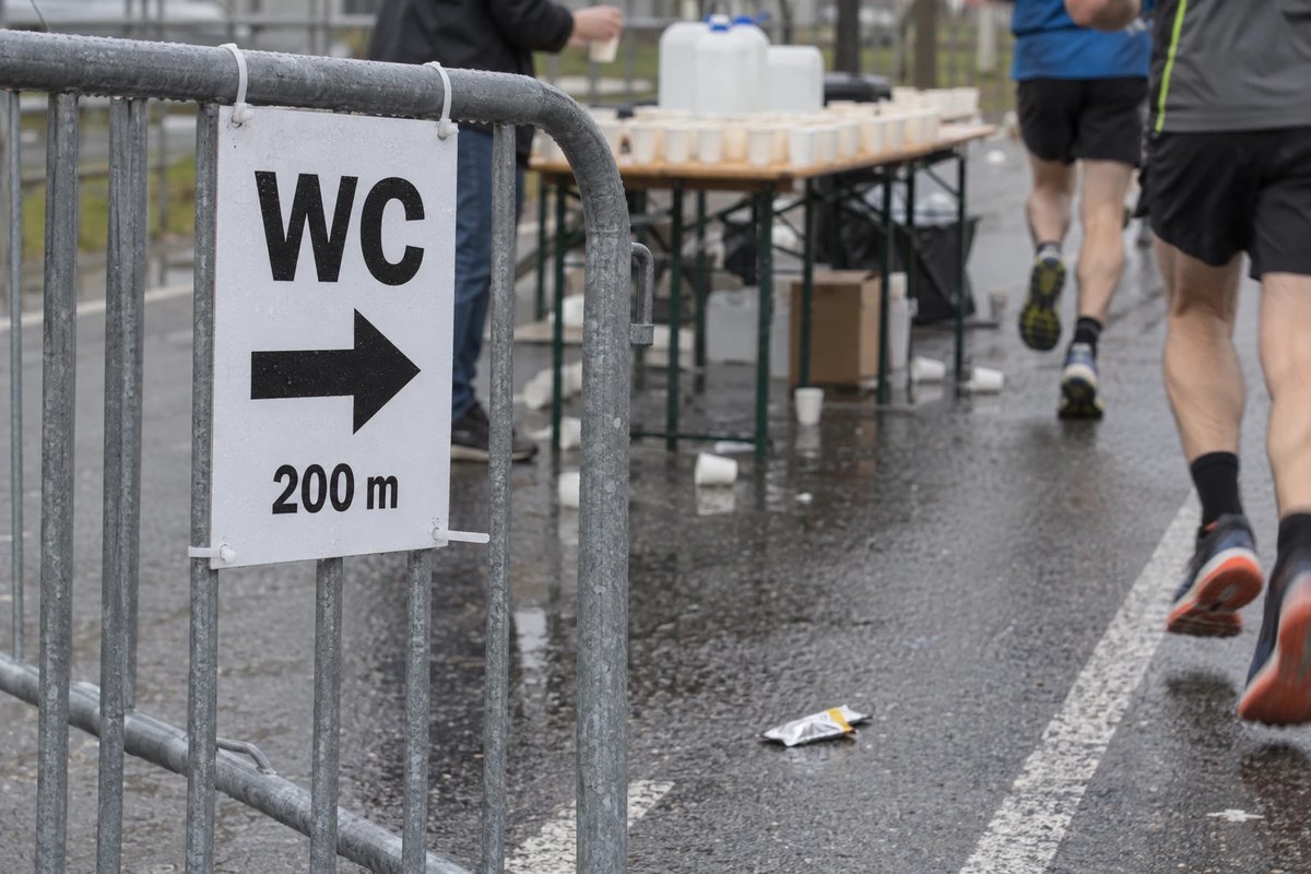 Schild mit der Aufschrift WC und einem Pfeil nach rechts, 200 m, an einem Absperrgitter neben einer Verpflegungsstelle mit Bechern, im Hintergrund laufen zwei Läufer auf nasser Straße vorbei