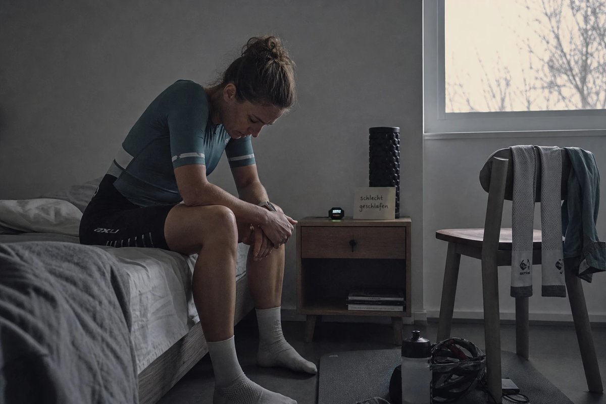 Erschöpfte Triathletin sitzt früh morgens auf der Bettkante vor dem Training, daneben eine Notiz über schlechten Schlaf