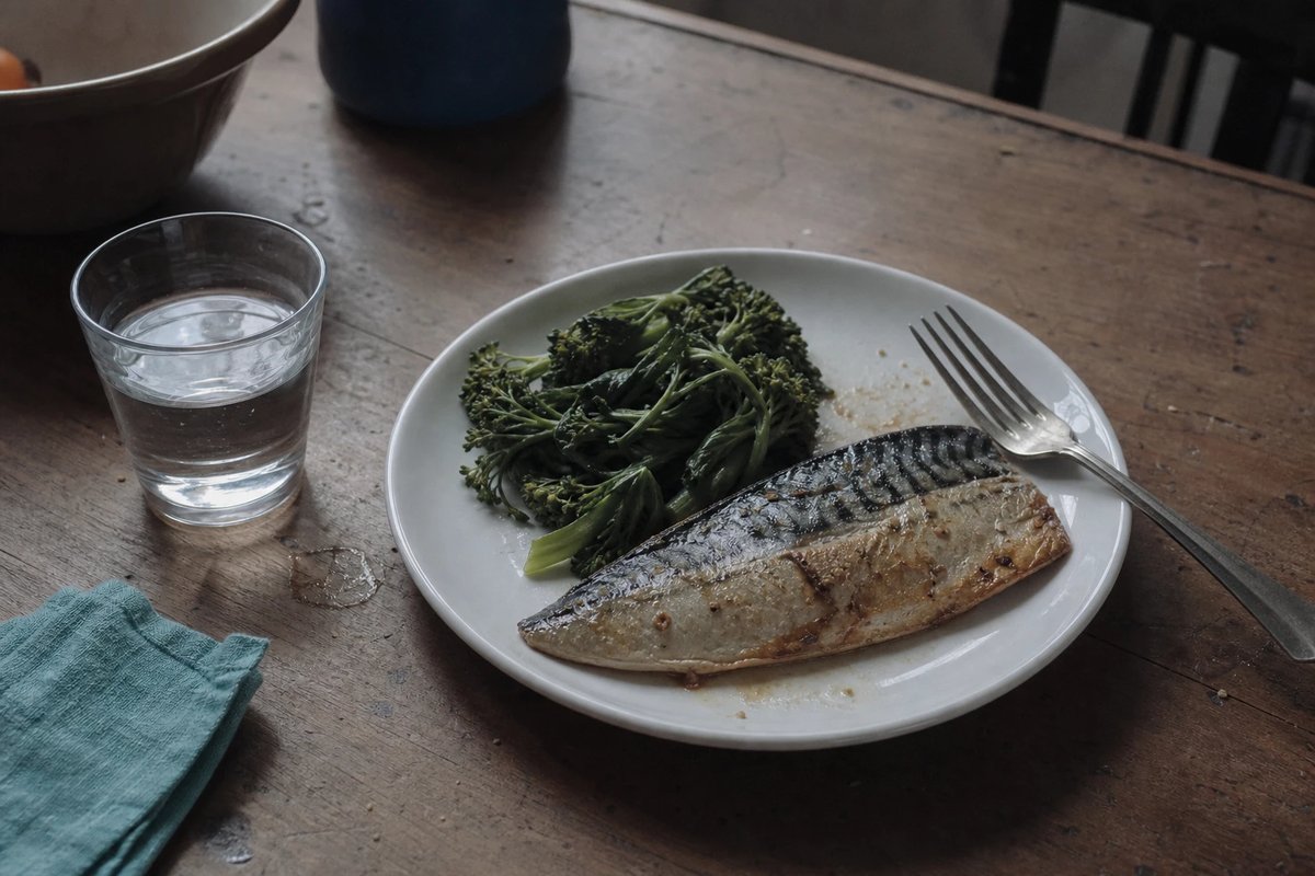 Teller mit gegartem Makrelenfilet und gedünstetem Grüngemüse auf einem Holztisch, daneben ein Glas Wasser und eine Gabel