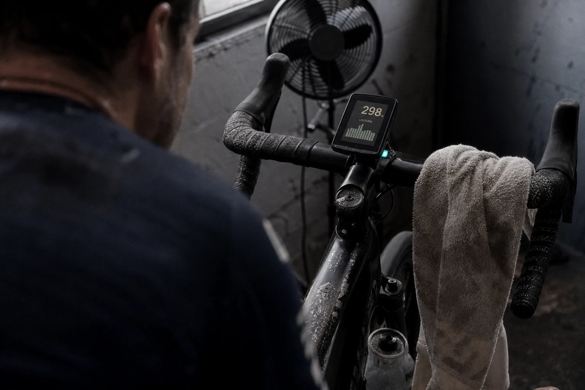 Über die Schulter des Fahrers fotografierter, am Vorbau montierter Radcomputer auf einem Rennrad am Smarttrainer im heimischen Pain Cave, das Display zeigt aufrecht zur Fahrerin gerichtet eine Wattzahl