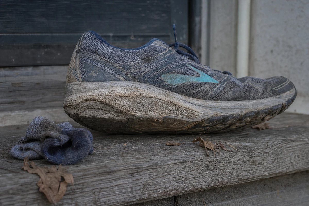Ein abgelaufener Straßenlaufschuh liegt seitlich auf einer verwitterten Holzschwelle, die äußere Kante der Sohle ist sichtbar stärker abgenutzt als der Rest