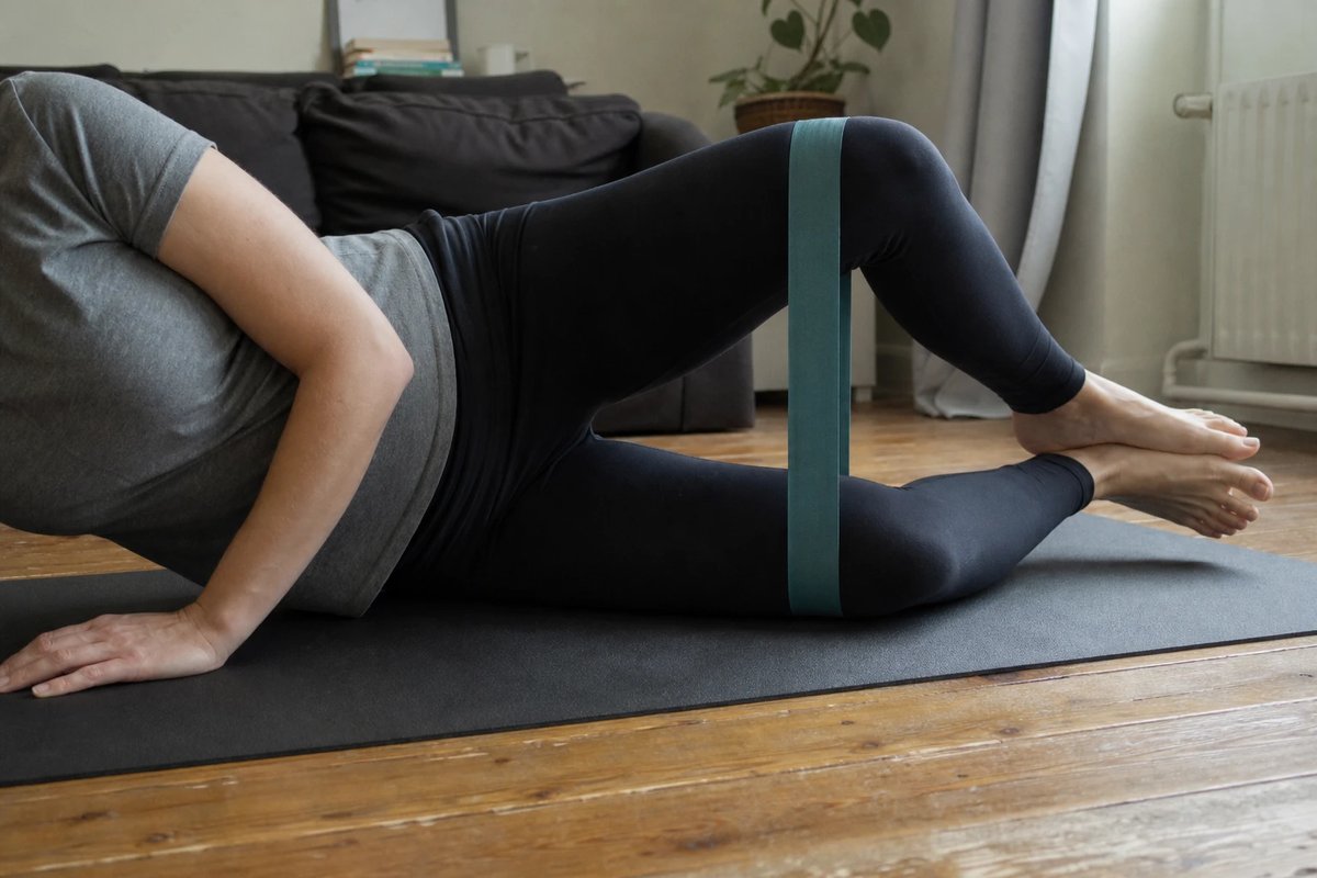 Person macht zu Hause auf einer Matte die Muschel-Übung in Seitlage, ein Widerstandsband um beide Oberschenkel, das obere Knie wird angehoben