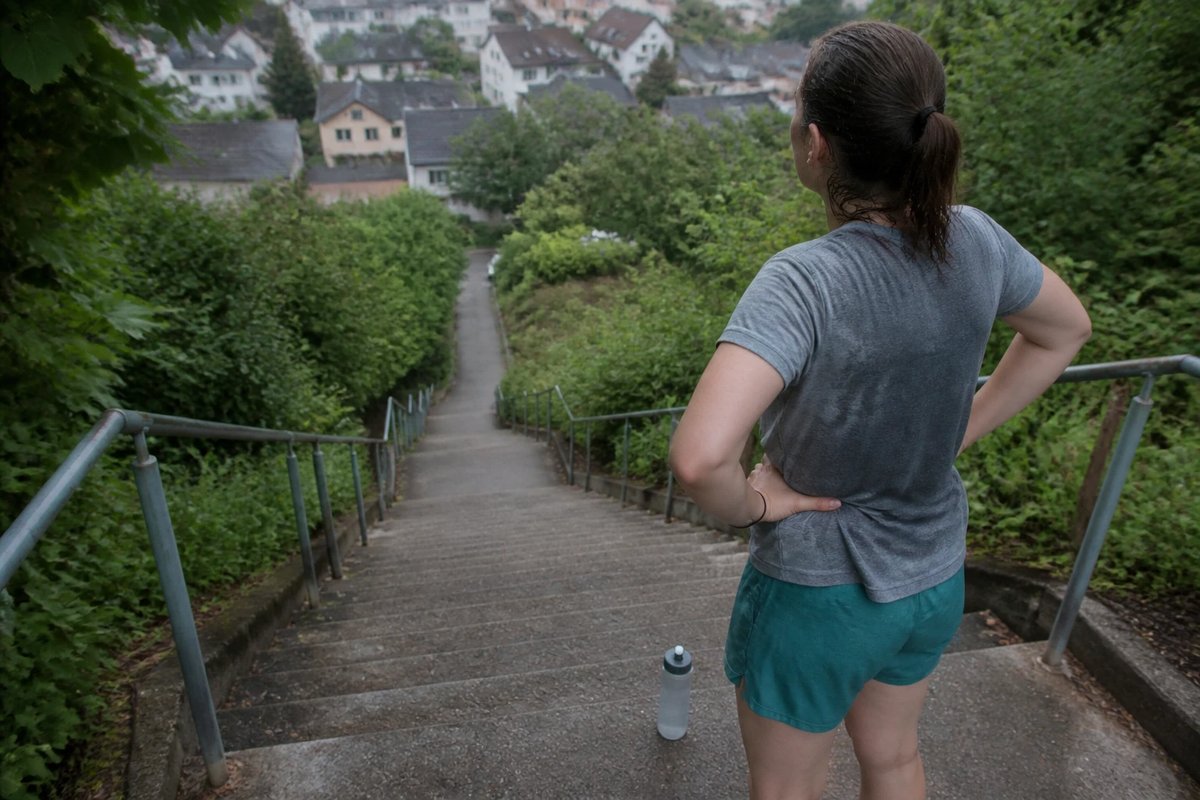 Eine Läuferin steht oben an einer langen Außentreppe, die Hände in die Hüften gestützt, und blickt zur Erholung zwischen den Intervallen die Stufen hinab