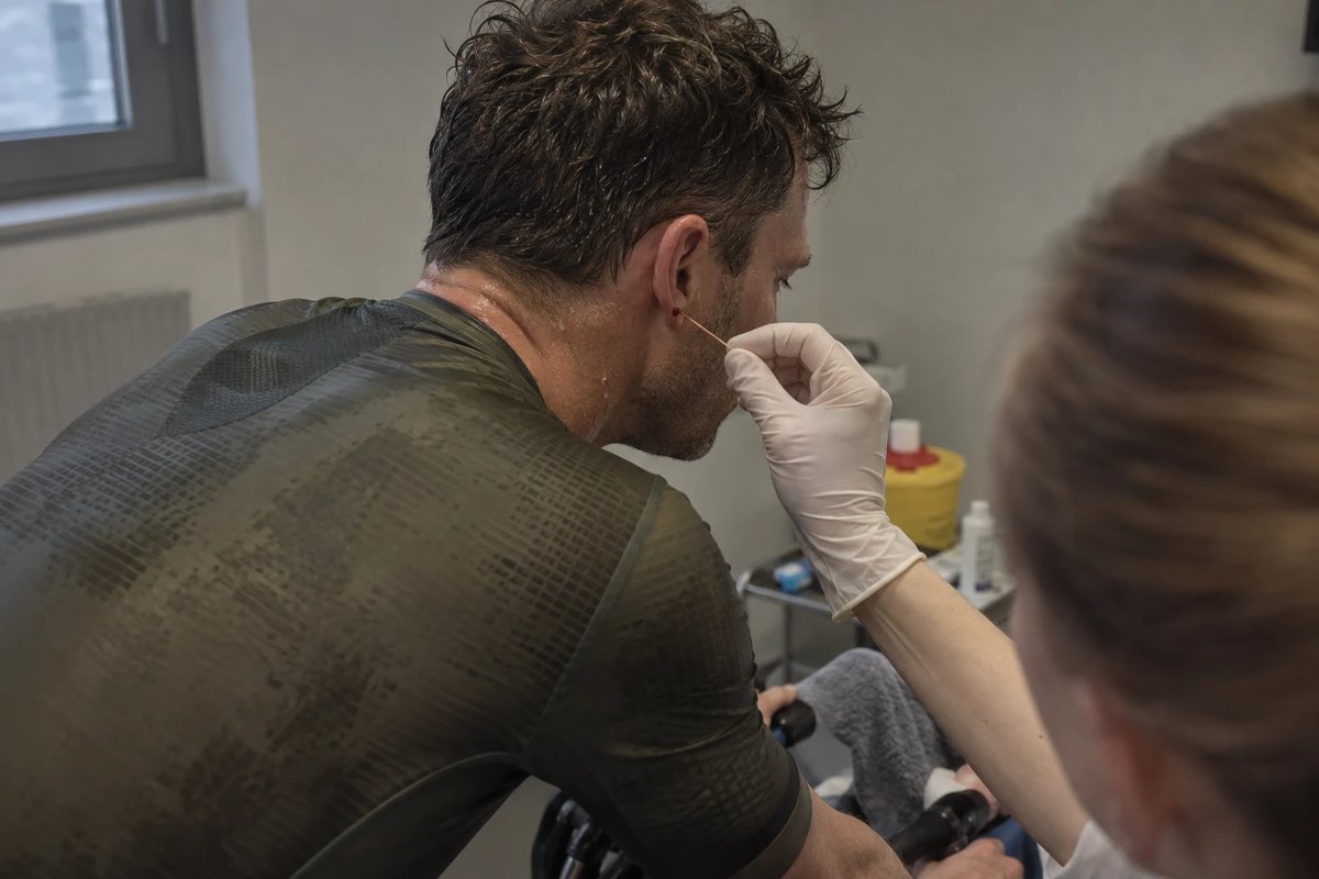 Radfahrer auf einem Rollentrainer während eines Dauertests, eine Hand mit Handschuh nimmt mit einer Kapillare Blut am Ohrläppchen ab