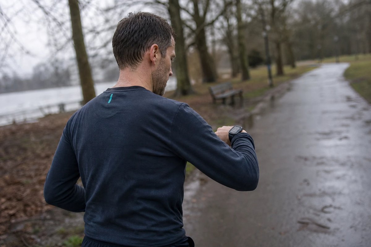 Läufer von hinten auf einem nassen Parkweg an einem grauen Morgen, der bei einem gleichmäßigen Tempolauf auf seine GPS-Uhr am Handgelenk blickt