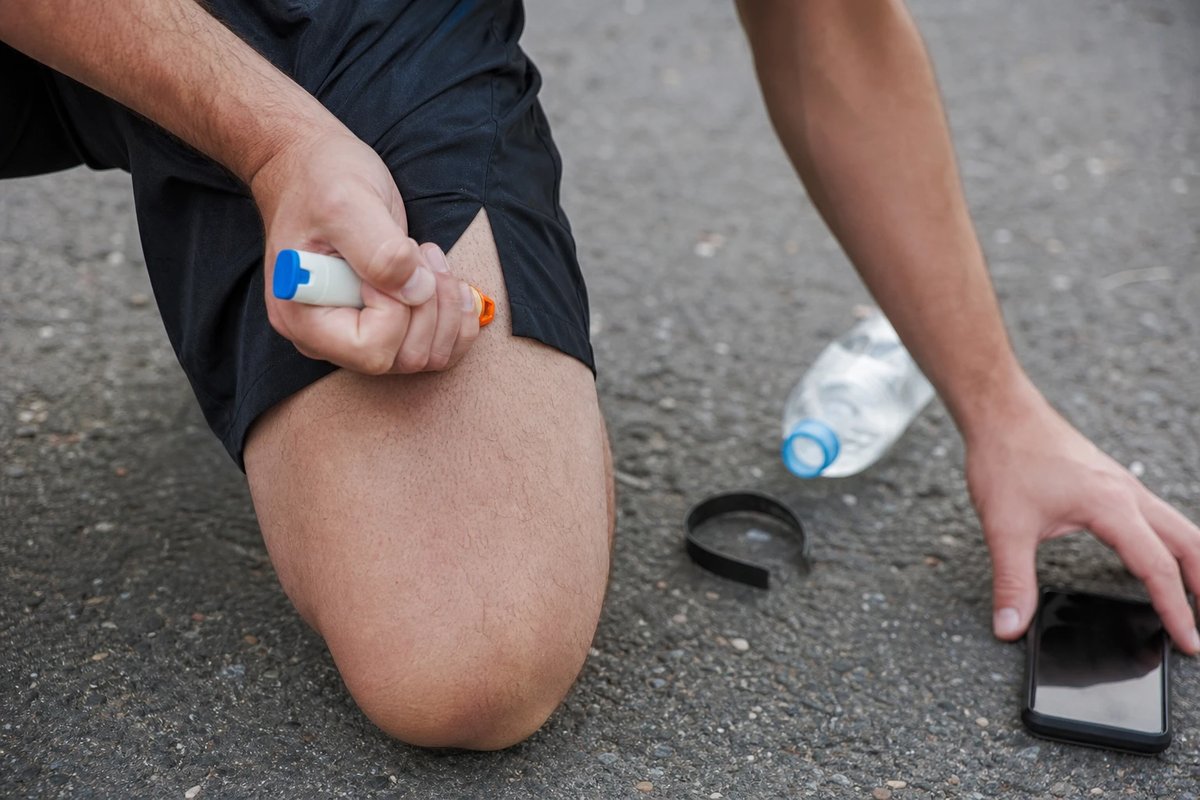 Hand drückt einen Adrenalin-Autoinjektor seitlich gegen den äußeren Oberschenkel durch die Laufhose, die andere Hand greift nach einem Handy auf dem Boden, daneben eine umgefallene Trinkflasche