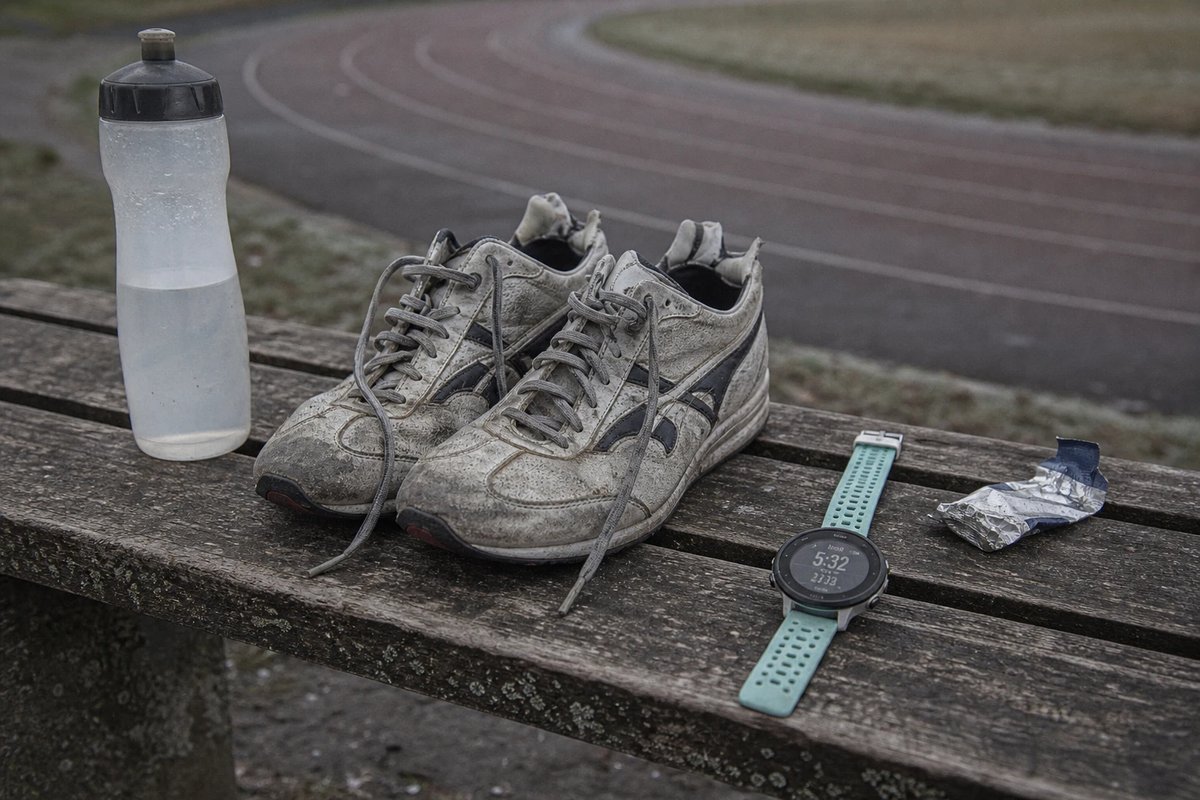 Gut eingelaufene leichte Wettkampfschuhe, eine GPS-Uhr mit teal Armband, eine halbleere Trinkflasche und ein zerknülltes Gel auf einer verwitterten Holzbank am Rand einer Laufbahn an einem grauen Morgen